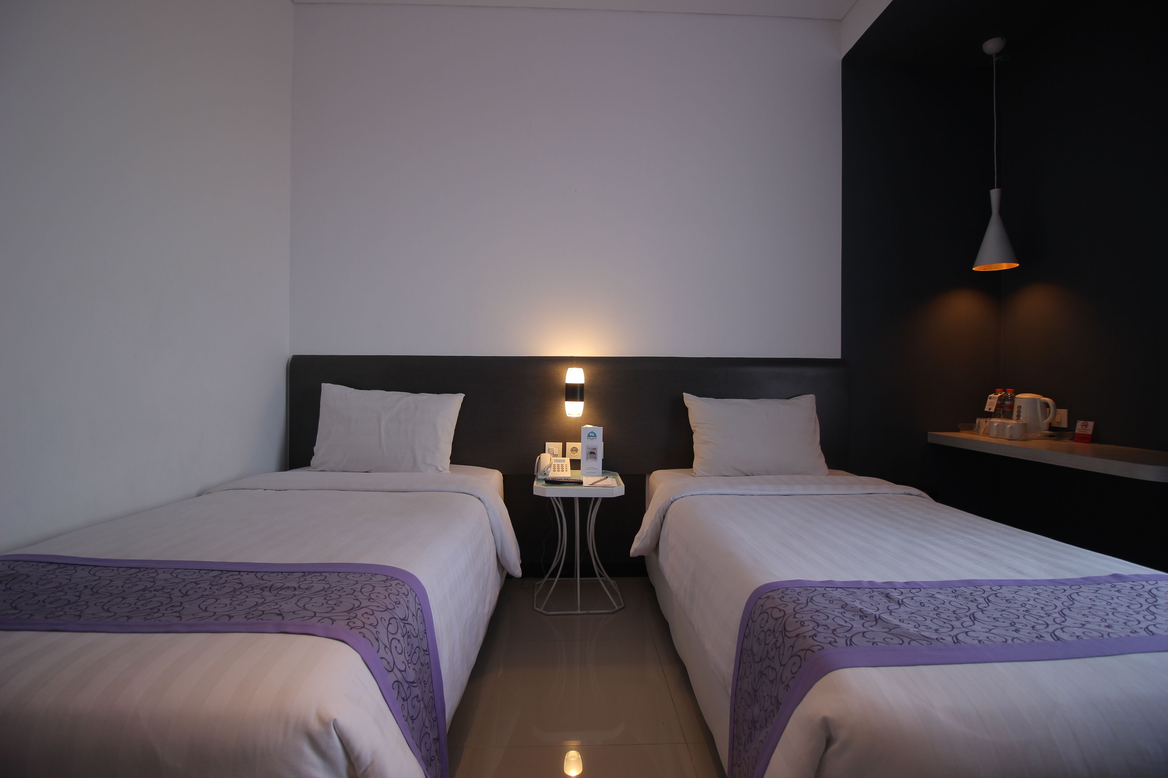 Een hotelkamer met twee bedden, een tafeltje met een telefoon en een lamp, en een plank met spullen.