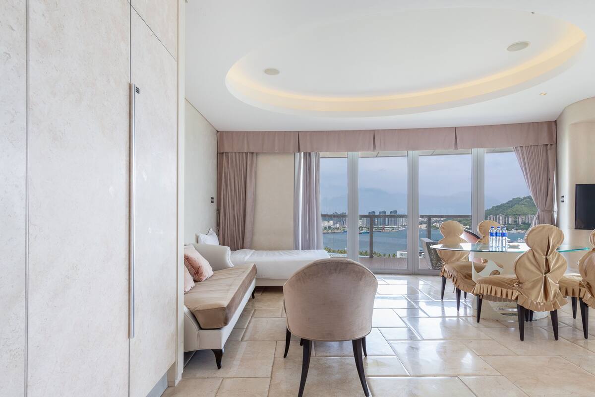 Habitació d'hotel moderna amb un llit gran, zona de seients, taula de menjador amb caires i vista a la ciutat a través de grans finestres.