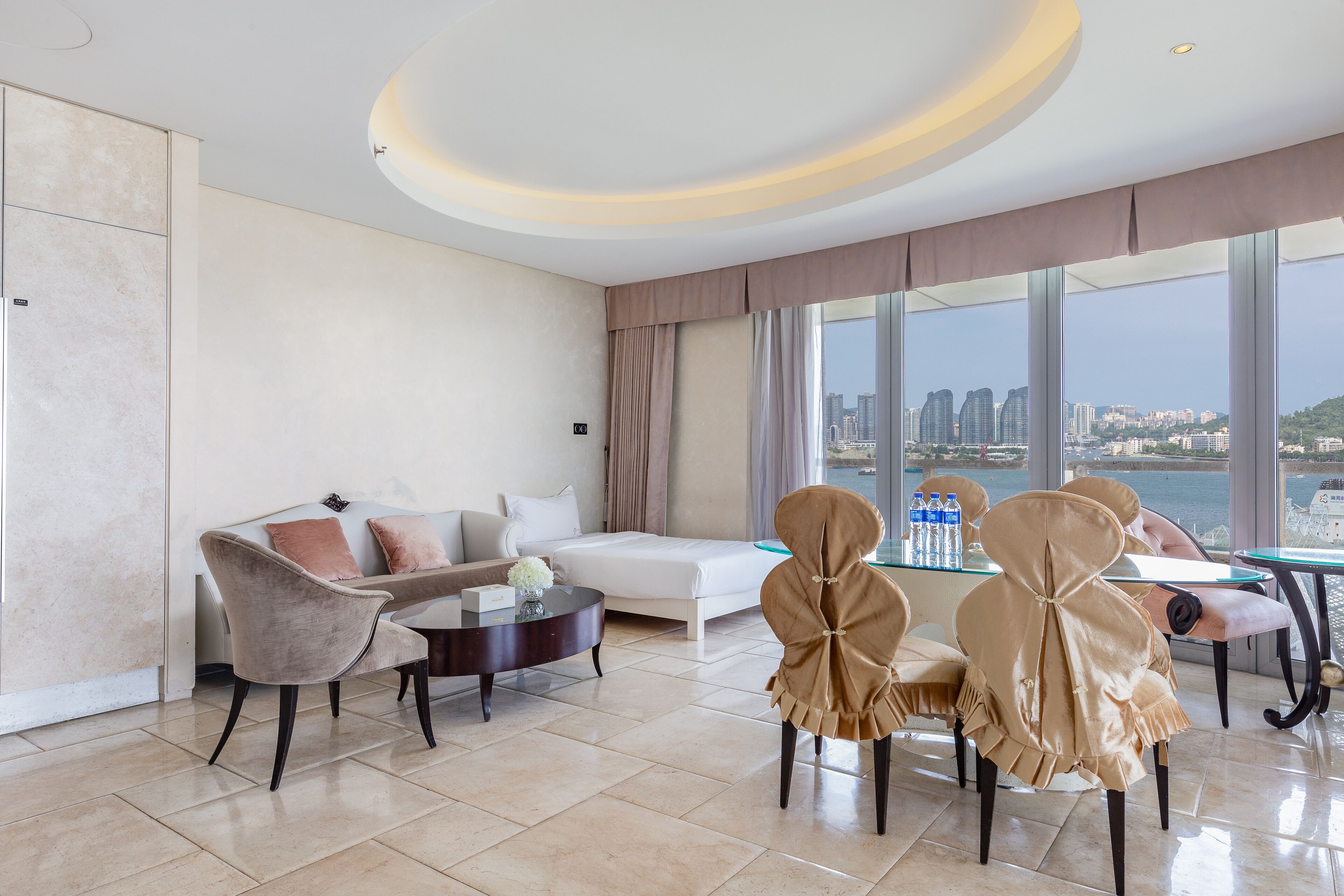 A modern hotel room with a sofa, dining table, chairs, and a large window offering a city view.