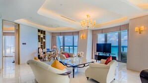 A modern living room with a glass coffee table, two sofas, a TV, and a large window offering a view of the sea.
