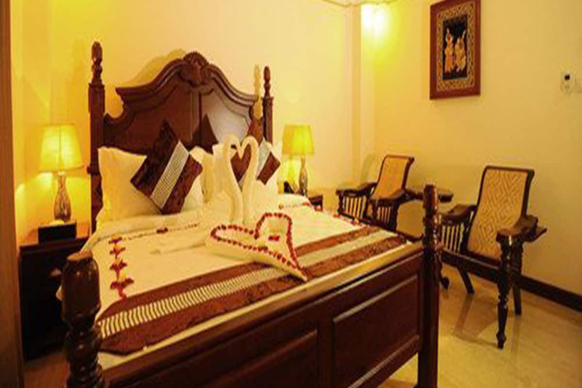 A neatly made bed with a heart-shaped pillow and a decorative pillow. There are two bedside tables with lamps, a chair, and a framed picture on the wall.