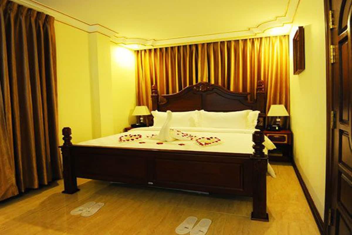 A neatly made bed with a heart-shaped arrangement of rose petals on the bedspread, flanked by two bedside tables with lamps, in a room with wooden flooring and curtains.