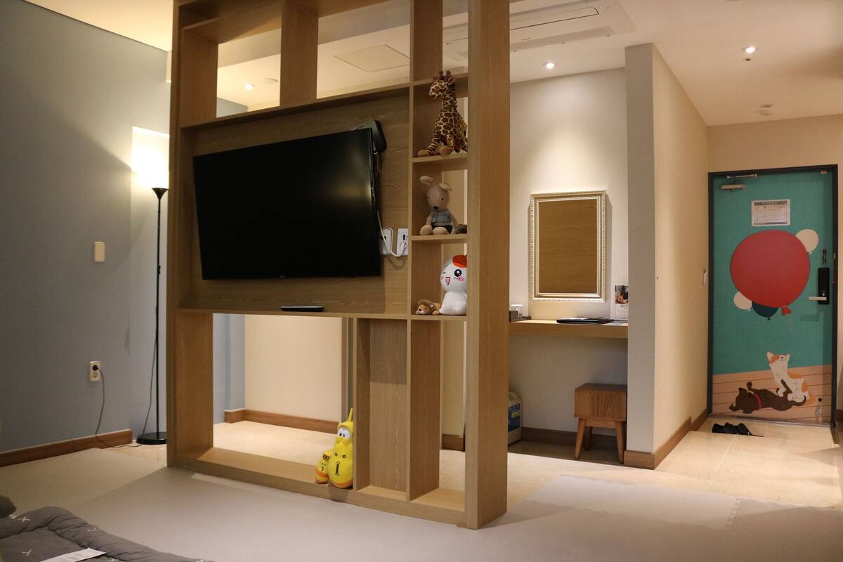 A room with a mounted television, wooden shelving with toys, a small stool, and a door with a cartoon mural.