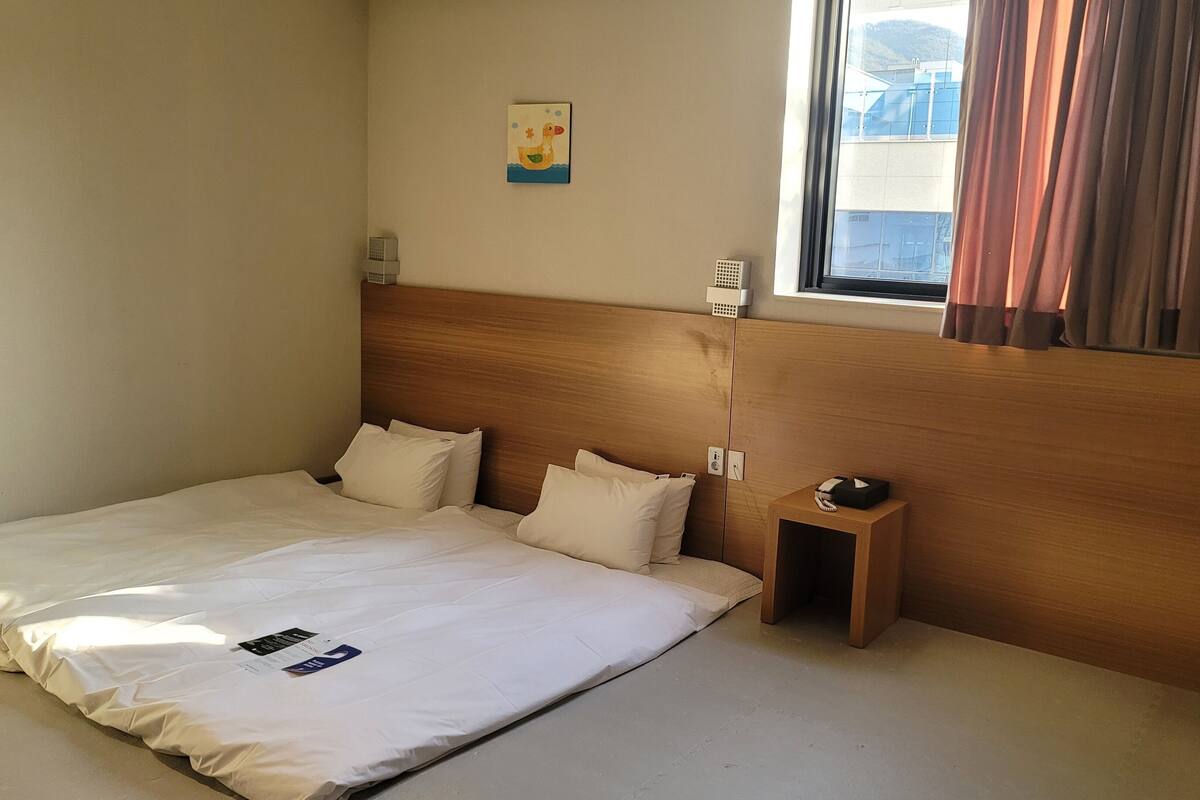 A hotel room with a wooden headboard, a single bed with white bedding, a small bedside table with a lamp, and a window with curtains.