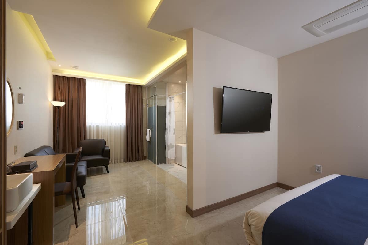 A hotel room with a bed, a desk with a chair, a television, and a bathroom visible through an open door.