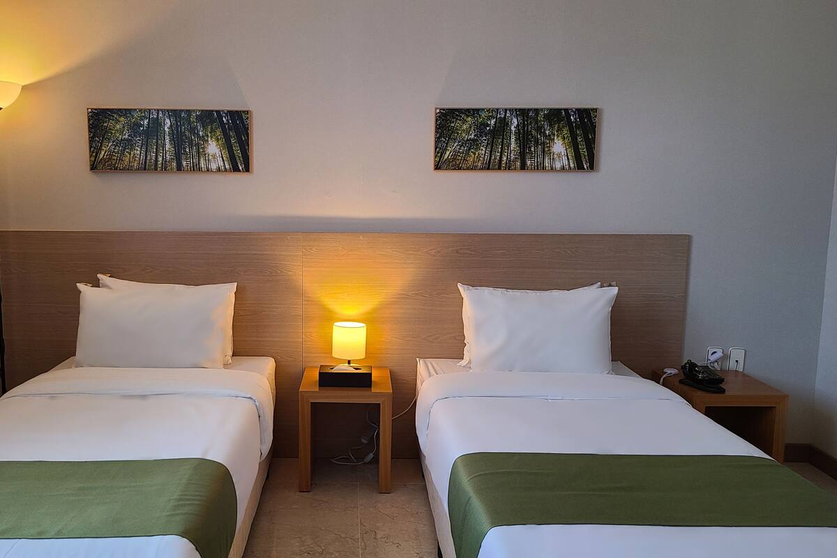 A hotel room with two beds, a nightstand with a lamp, and two wall-mounted paintings.