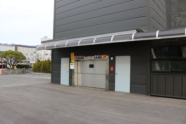 A building with a large garage door, a signboard with various symbols, and a parking lot with a tree and a stone wall. - Benikea Yangsan Hotel (Yangsan)