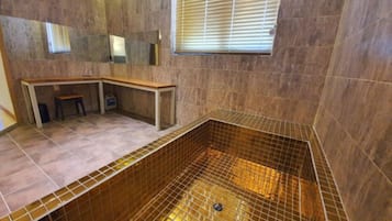 Un baño con suelo de baldosas, un banco, zona de ducha con rejilla metálica y una ventana con persianas.