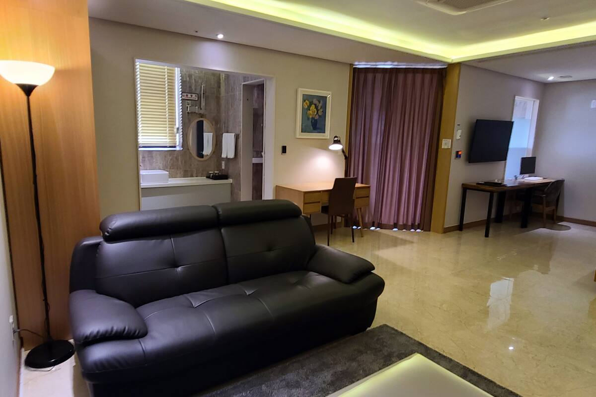 A modern hotel room with a black leather sofa, a desk with a lamp, a bed, a TV, and a bathroom with a mirror and sink.