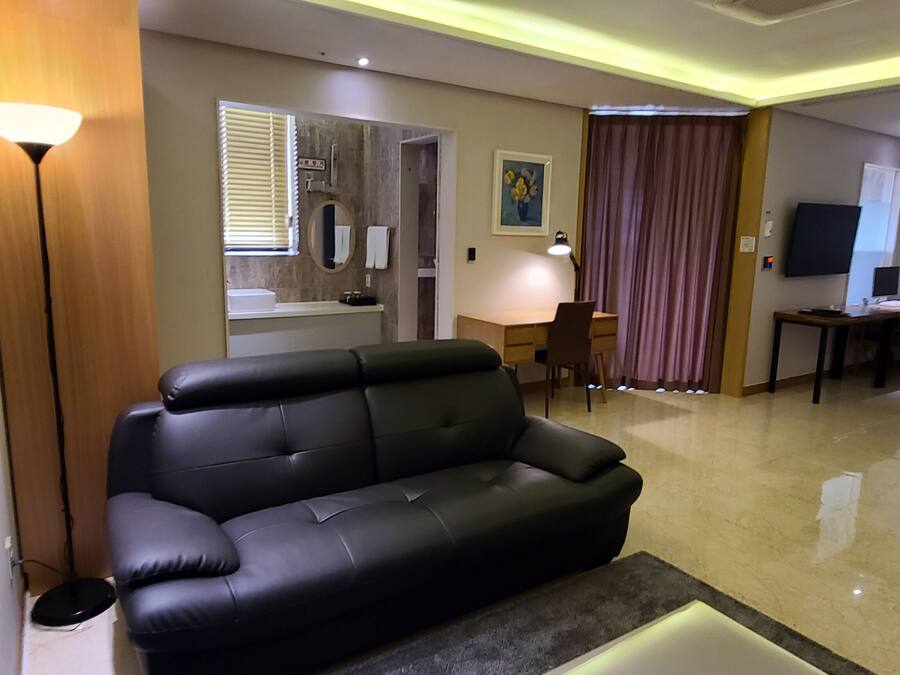 Una habitación de hotel moderna con un sofá de cuero negro, un escritorio con lámpara, una cama, un televisor y un baño con espejo y lavamanos.