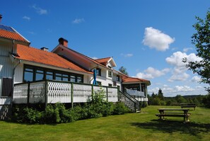 Un bâtiment à deux étages, avec un toit recouvert de tuiles rouges, une façade blanche et un balcon. On aperçoit des tables de pique-nique au premier plan.