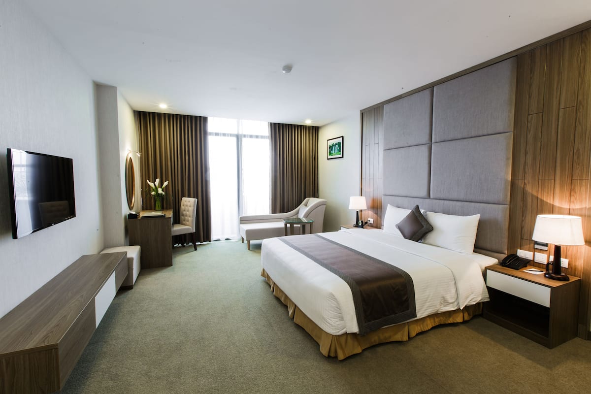 Habitación de hotel con una cama grande, una mesita de noche con lámpara, una silla, un escritorio, un televisor de pantalla plana montado en la pared y una ventana con cortinas.