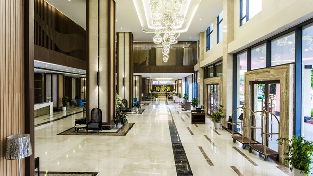 A spacious hotel lobby with a high ceiling, a central chandelier, and a reception desk.