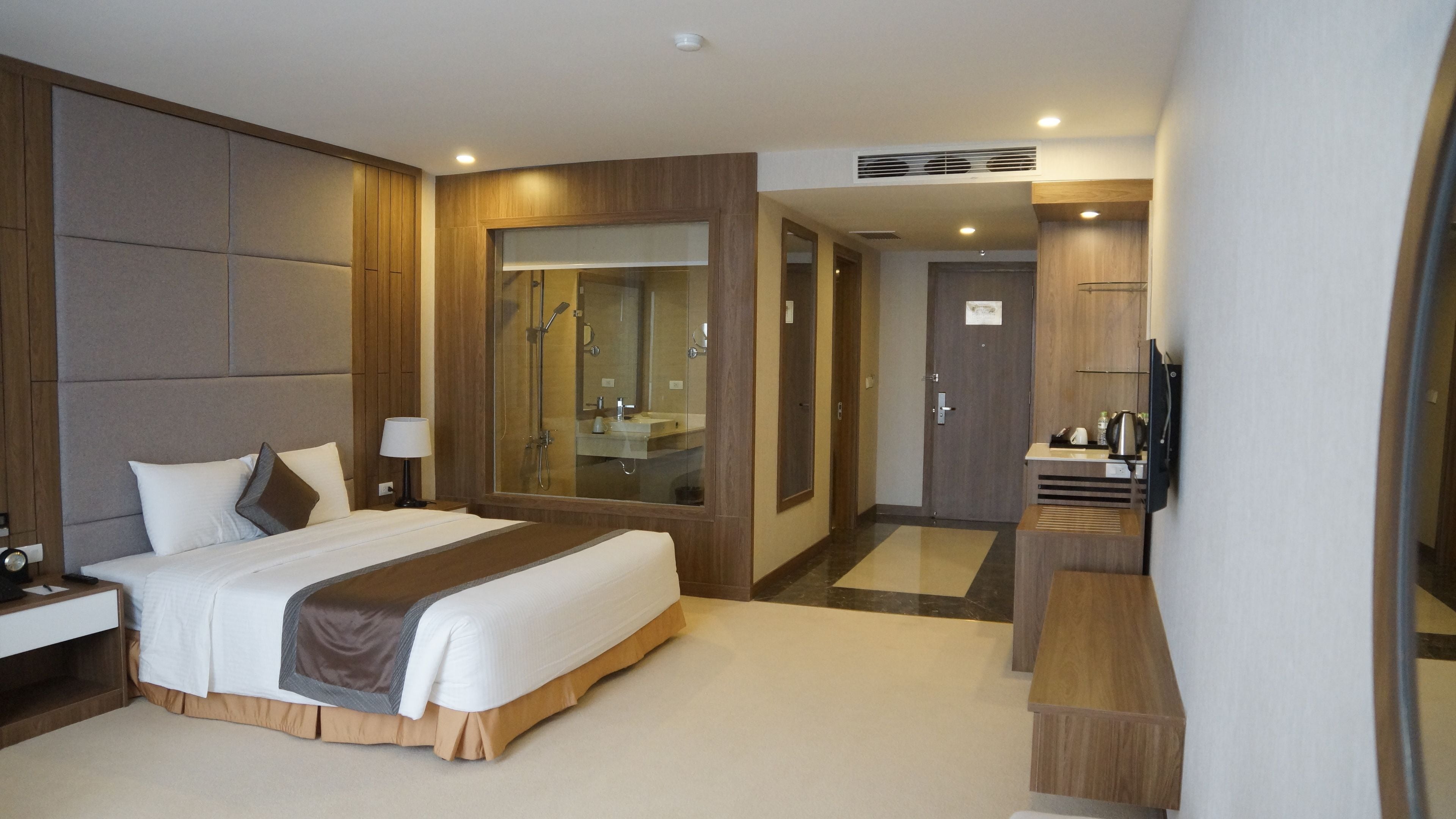 A modern hotel room with a large bed, a bedside table, a mirror, a shower area, and a small desk with a chair.