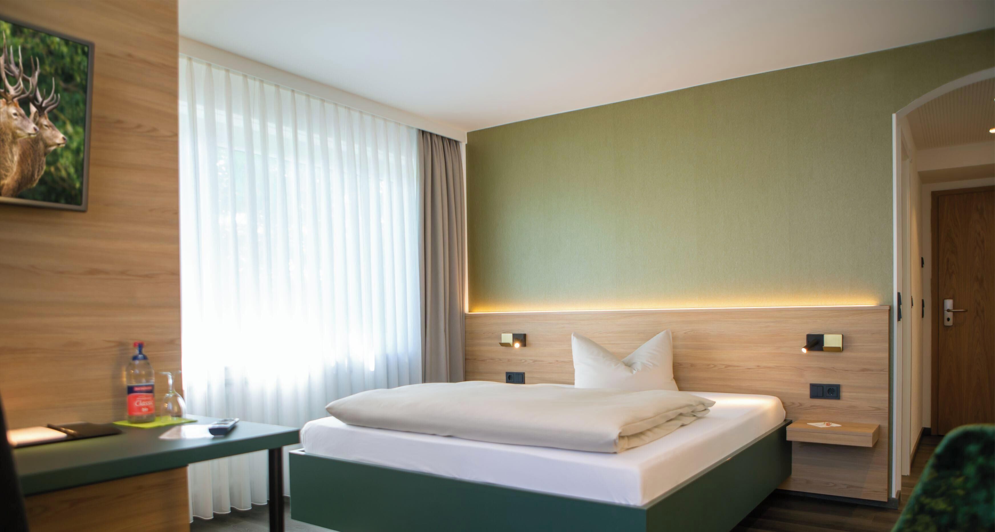 Hotellihuone, jossa on sänky, yöpöydät, yölamppu, seinään kiinnitetty valaisin ja ikkuna, jossa on verhot.