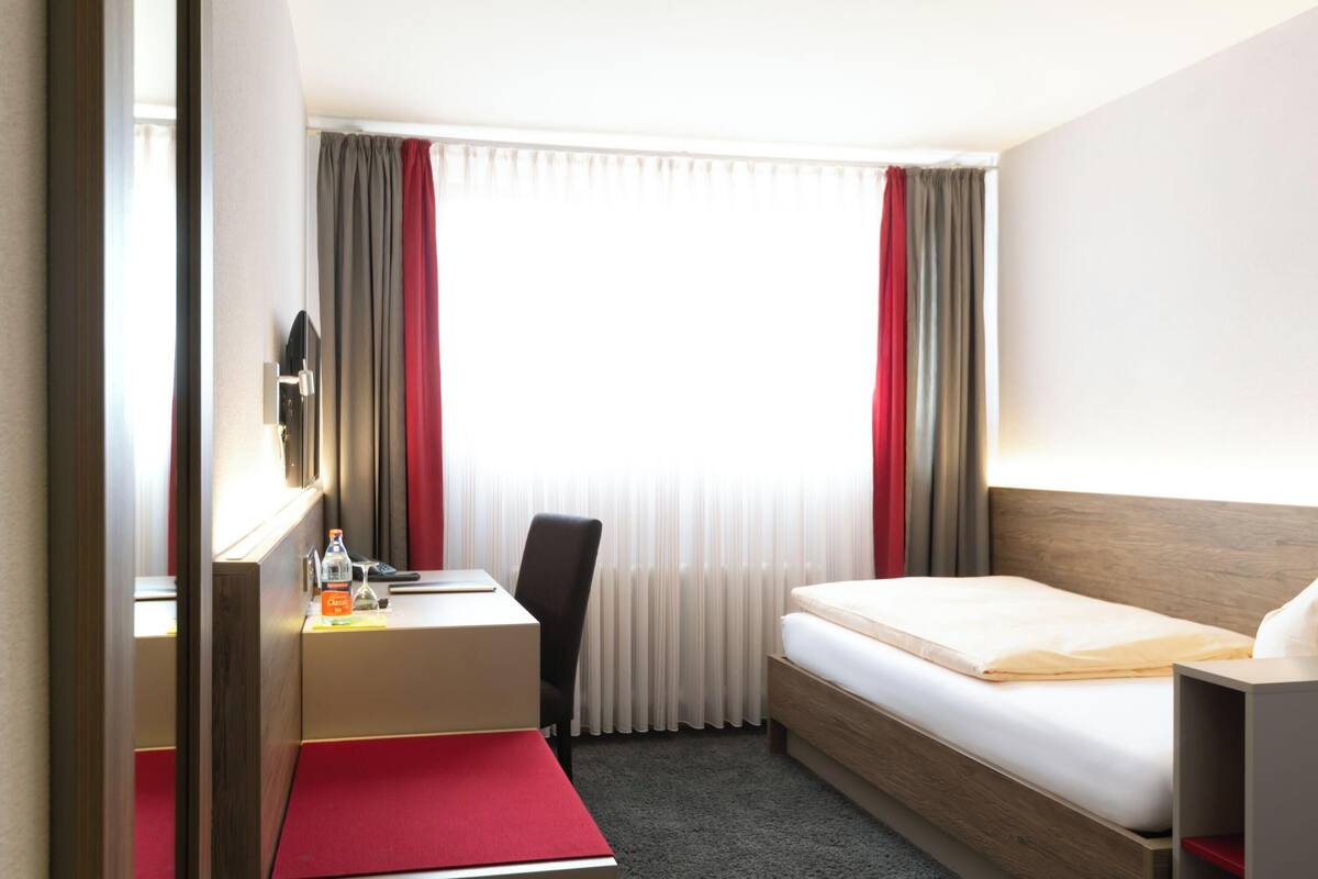 Ein Hotelzimmer mit einem Bett, einem Schreibtisch, einem Stuhl, einem Spiegel und einem Vorhangfenster.