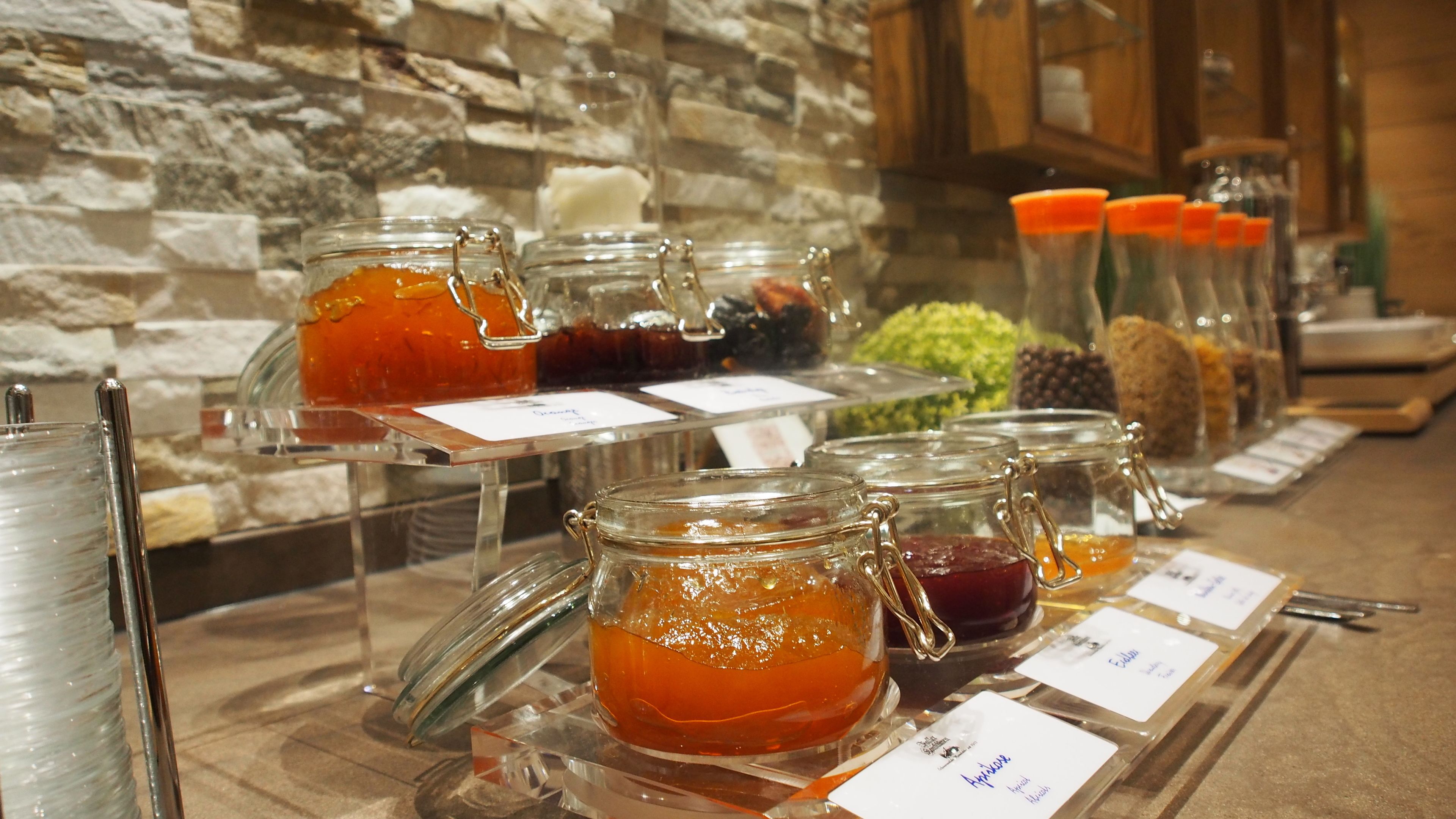 A buffet with various food items in glass jars, including jams and preserves, displayed on a counter with labels.