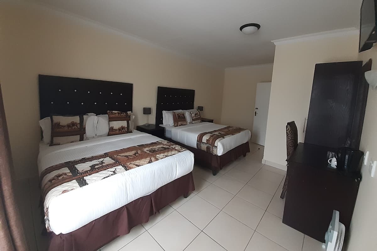 A hotel room with two beds, each with a headboard and pillows, a flat-screen TV mounted on the wall, and a small table with a lamp.