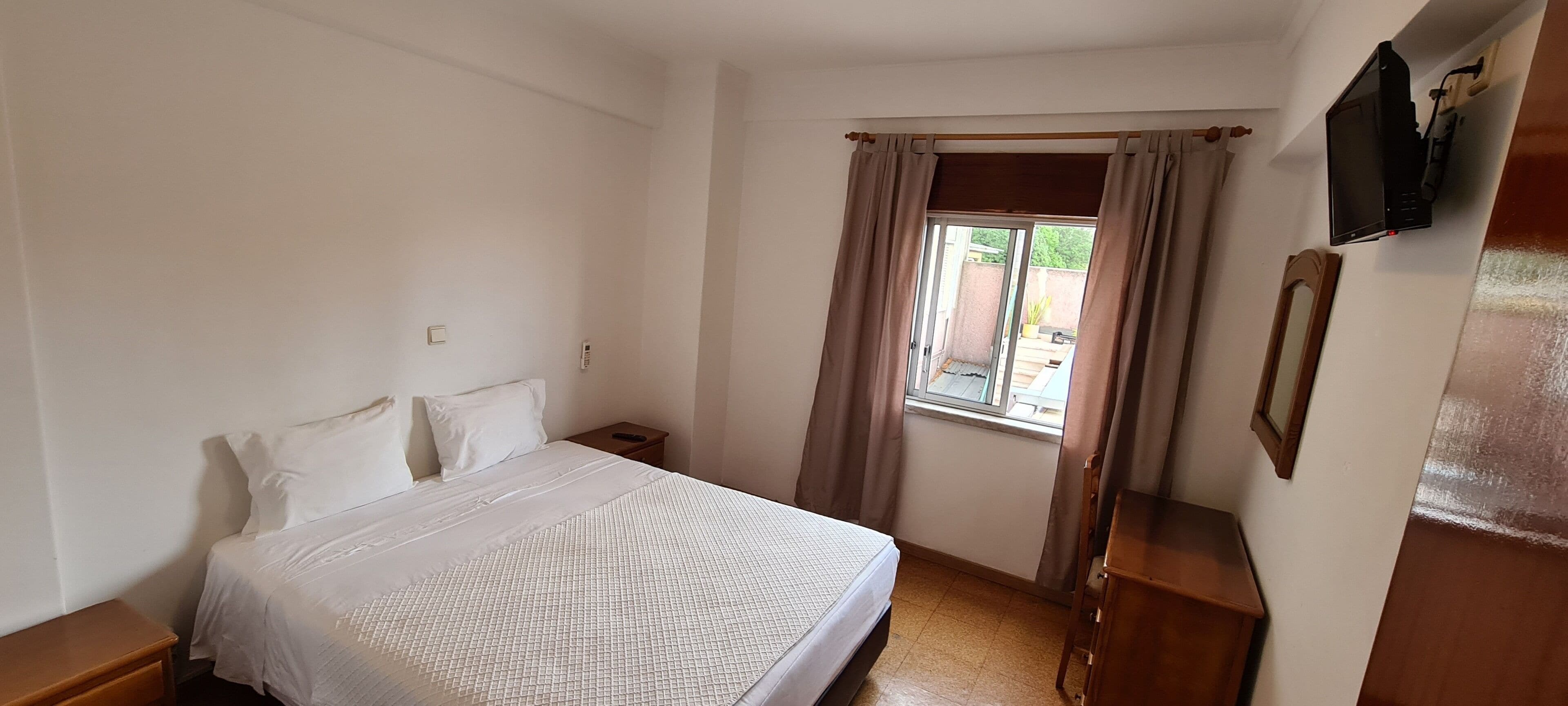 Un dormitorio con cama, mesita de noche, televisor montado en la pared y ventana con cortinas.