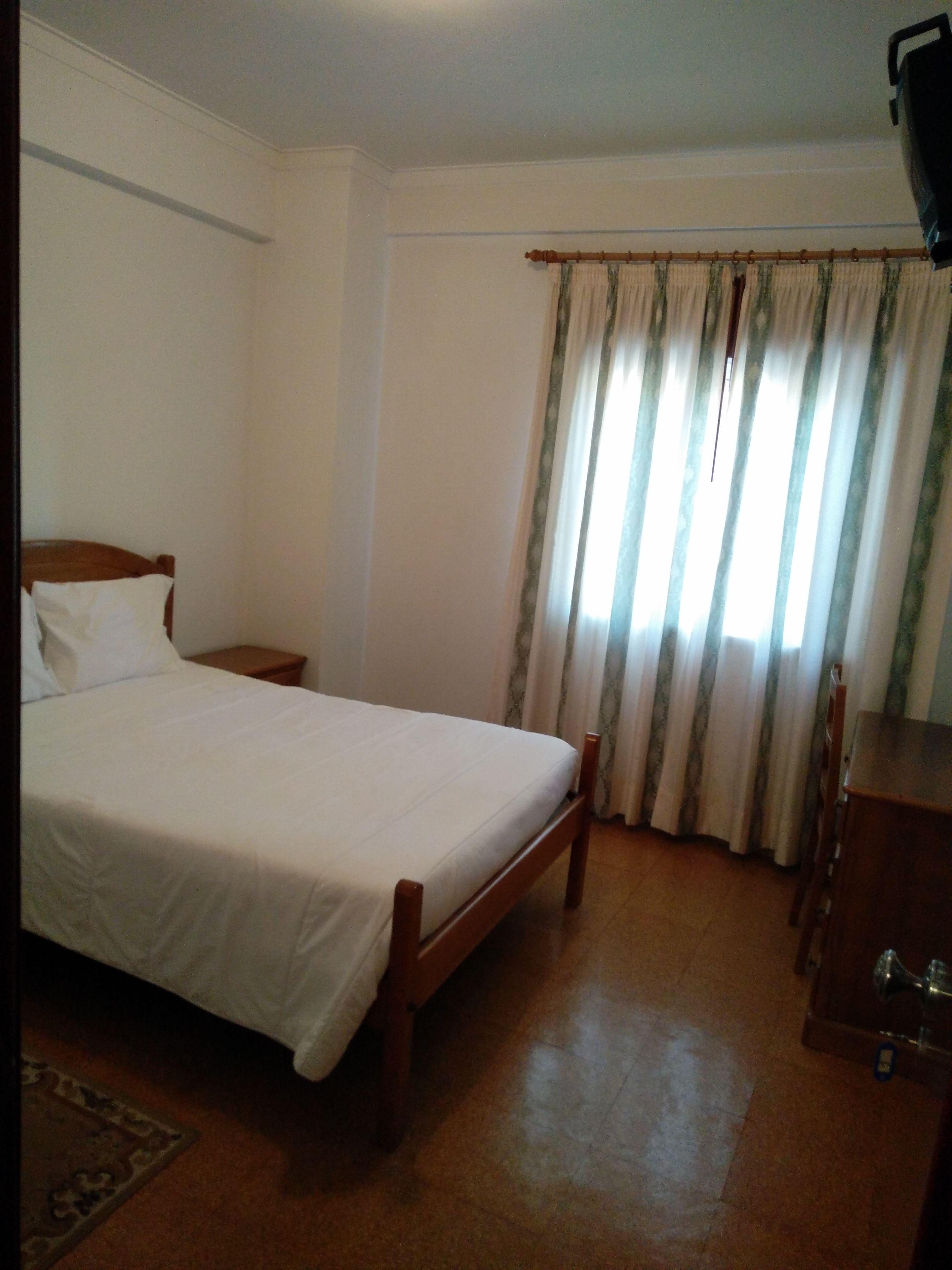 Un dormitorio con cama, una mesita de noche de madera, una silla, un escritorio y una ventana con cortinas.