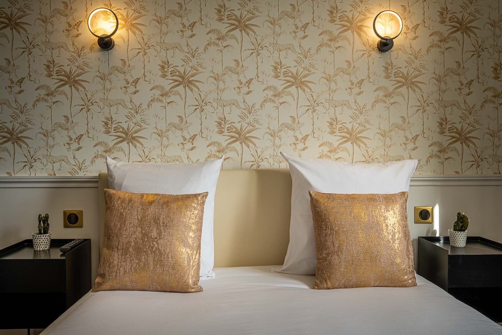 A hotel room with a bed, two gold-textured pillows, two nightstands with decorative items, and a wall with a floral pattern.