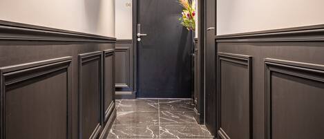 A hallway with dark wooden paneling, marble flooring, framed pictures on the walls, and an exit door with a flower arrangement.