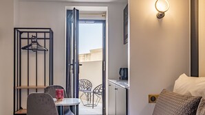 A hotel room with a bed, a small table with a chair, a hanging coat rack, and a view of the outside balcony. - Mijo's Appartements Hôtel (Fontenay-sous-Bois)
