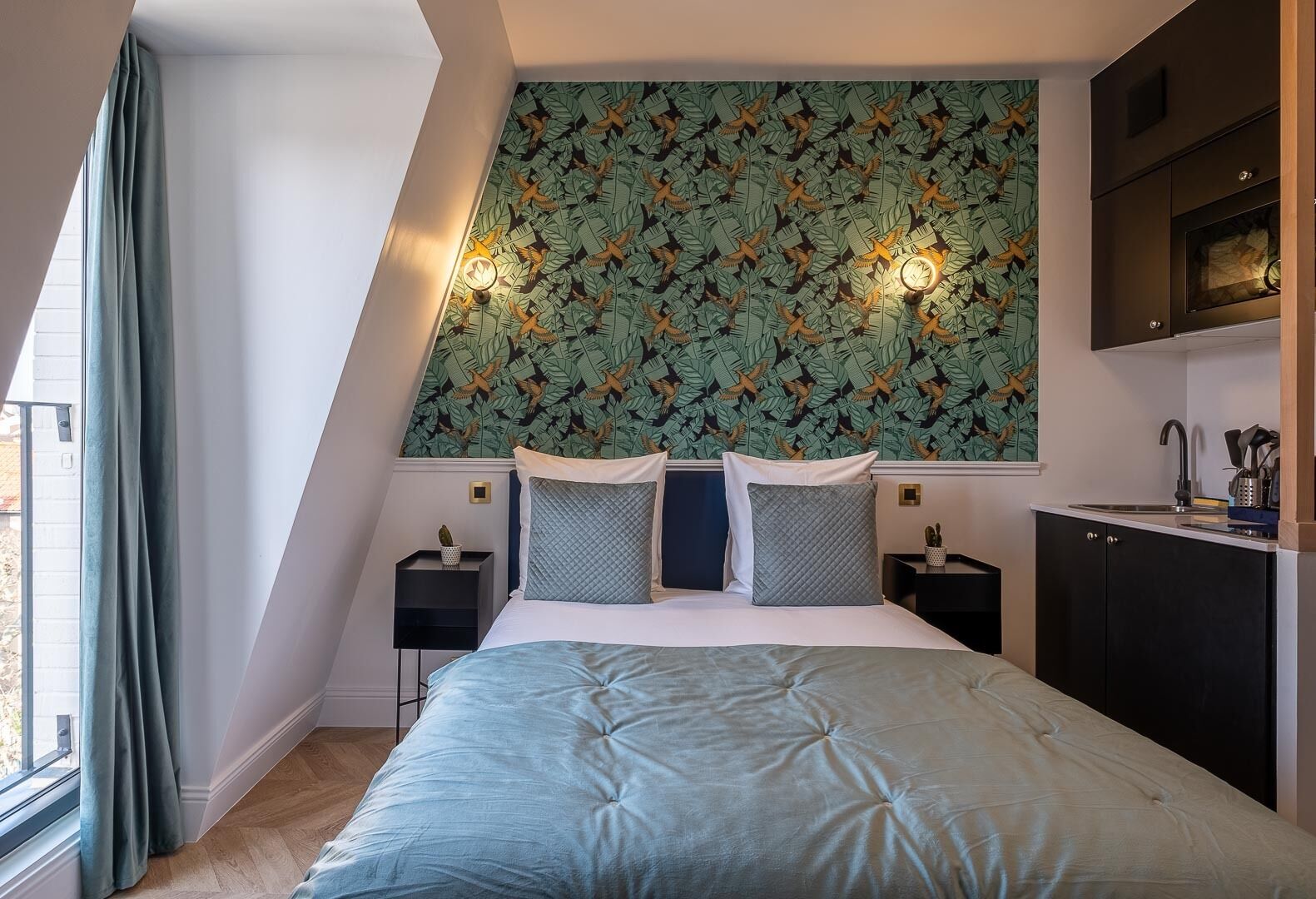 A bedroom with a bed, two bedside tables, a wall-mounted light, a window with curtains, and a kitchen area with a sink and cabinets.
