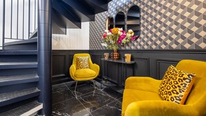 A staircase with black railings and a black and white patterned wall. There are two yellow armchairs with leopard print pillows, a small table with decorative items, and a vase with flowers. - Mijo's Appartements Hôtel (Fontenay-sous-Bois)