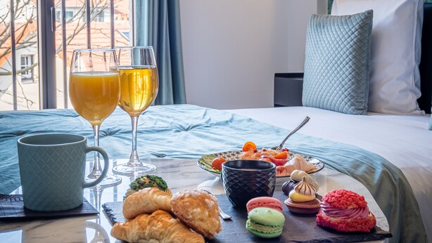 A neatly arranged breakfast table with croissants, macarons, and a fruit plate, set on a bed with a view of buildings through a window. - Mijo's Appartements Hôtel (Fontenay-sous-Bois)