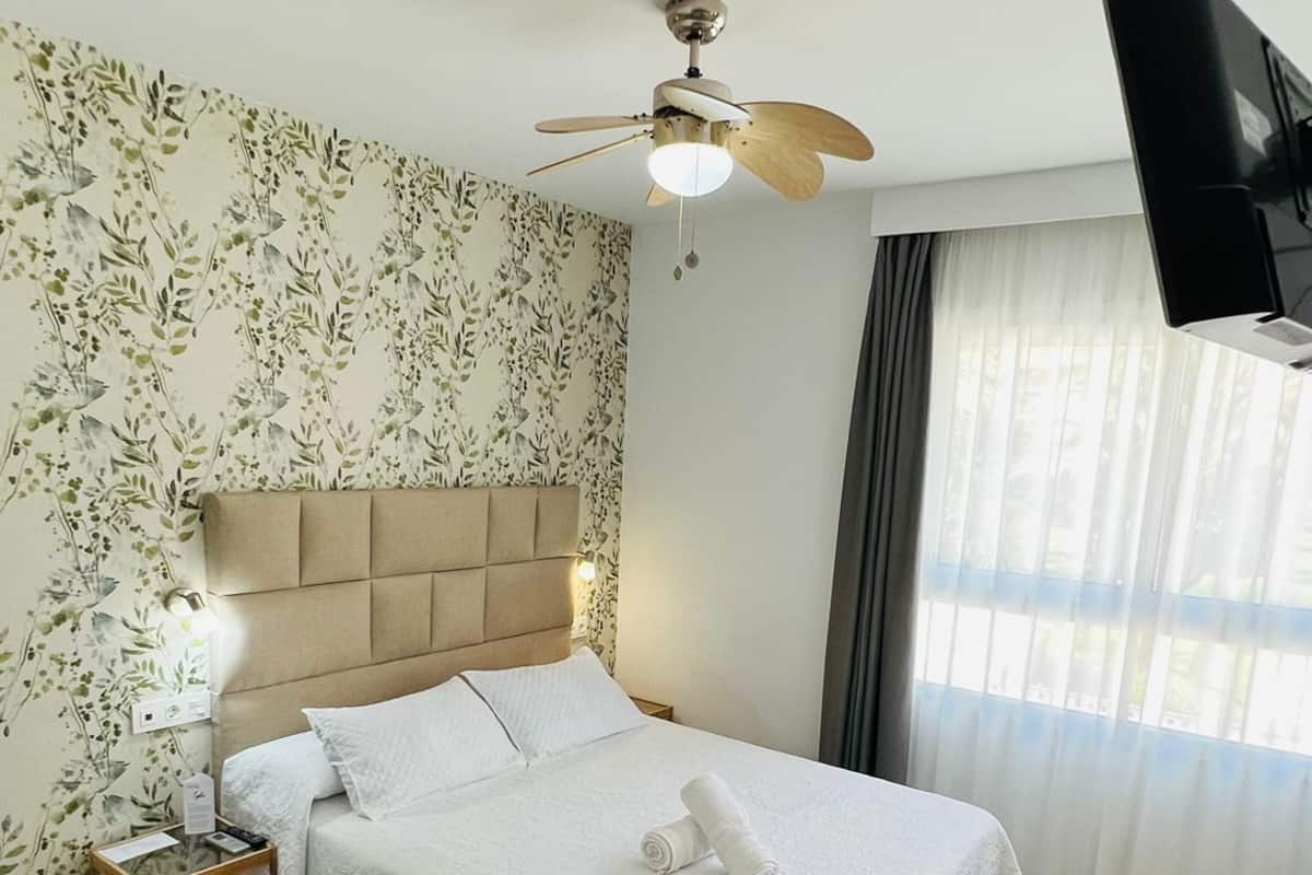 A hotel room with a bed, a desk, a chair, a ceiling fan, and a TV mounted on the wall.