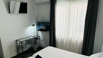 Quarto de hotel com duas camas, um ventilador de teto, televisão de tela plana, ar condicionado, uma mesa com cadeira e uma janela com cortinas.