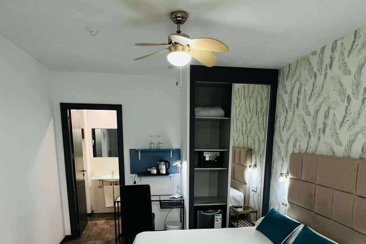 A bedroom with a large bed, a ceiling fan, a desk with a chair, a wardrobe, and a bathroom visible through an open door.