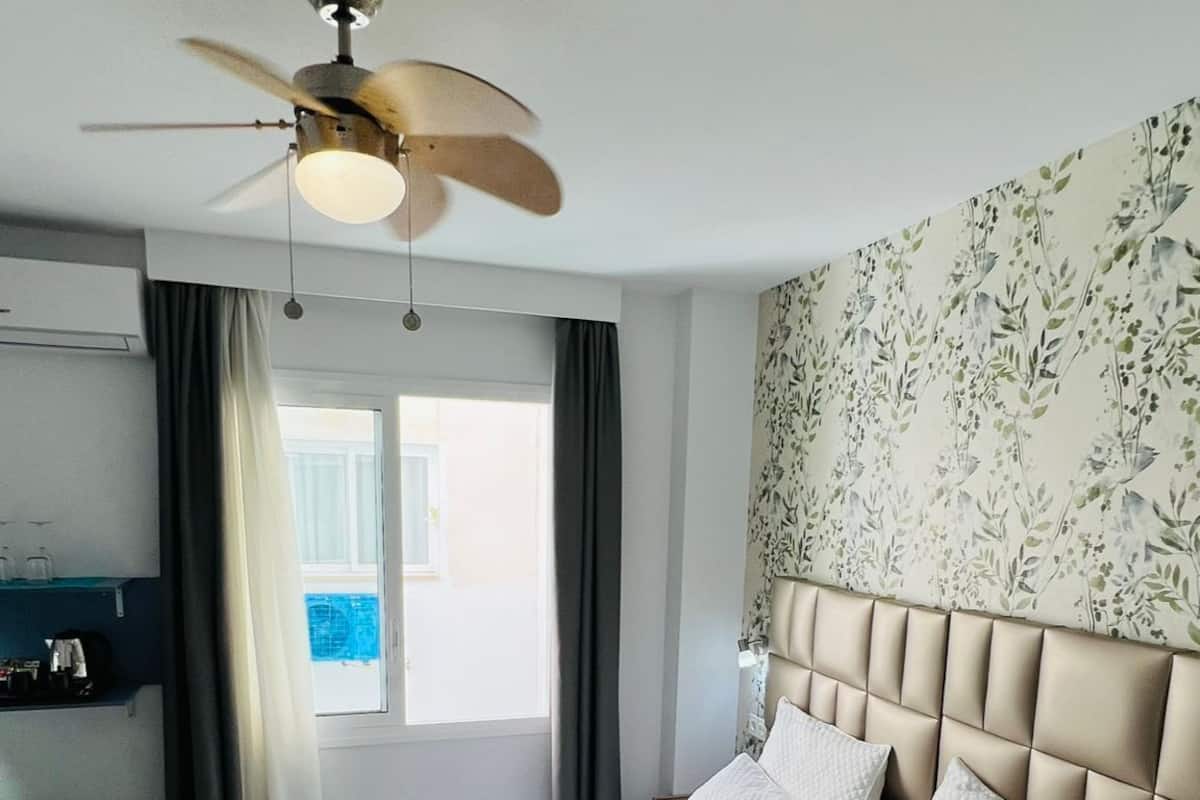 A hotel room with a bed, a ceiling fan, a window with curtains, and a wall with floral wallpaper.