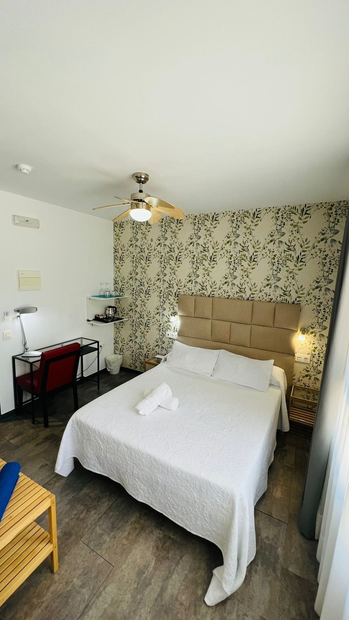 A hotel room with a bed, a desk with a chair, a ceiling fan, and floral wallpaper.