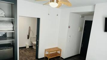 Habitación de hotel con cama, ventilador de techo, un taburete pequeño de madera, un armario y un baño visible a través de una puerta abierta.