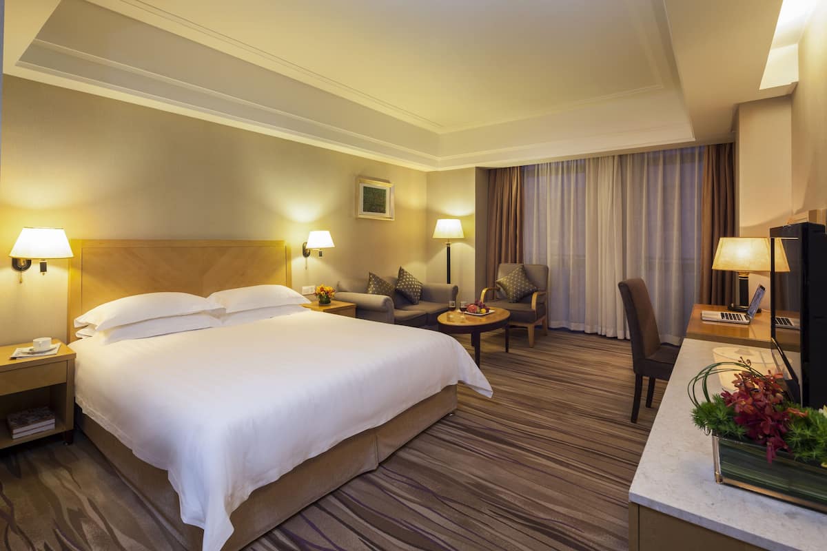 A hotel room with a large bed, a desk with a laptop, a chair, a sofa, and a small table with a vase of flowers.