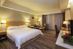 A hotel room with a large bed, a desk with a laptop, a chair, a sofa, and a small table with a vase of flowers. - Highton International (Shanghai)