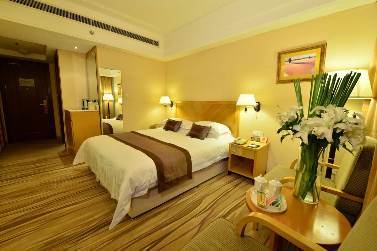 A hotel room with a large bed, bedside tables, a desk, a chair, a mirror, and a vase with flowers.