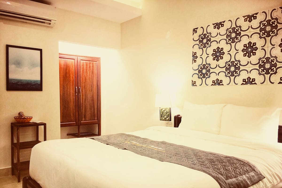 A neatly made bed with a patterned throw, a wooden wardrobe, a small table with a bowl, and a wall-mounted lamp.