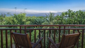 Un balcon avec deux chaises en bois donnant sur un paysage verdoyant, parsemé de palmiers, et offrant une vue sur l’océan.