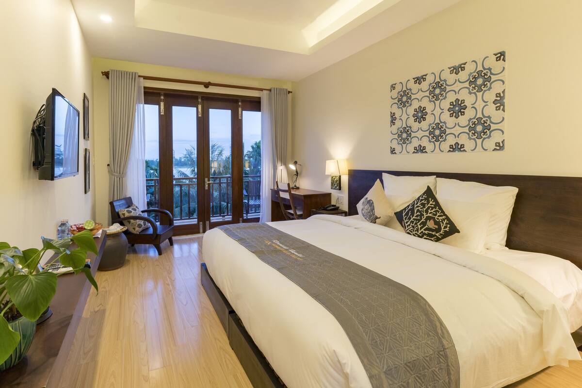 A hotel room with a large bed, a desk with a chair, a TV, and a balcony with curtains.