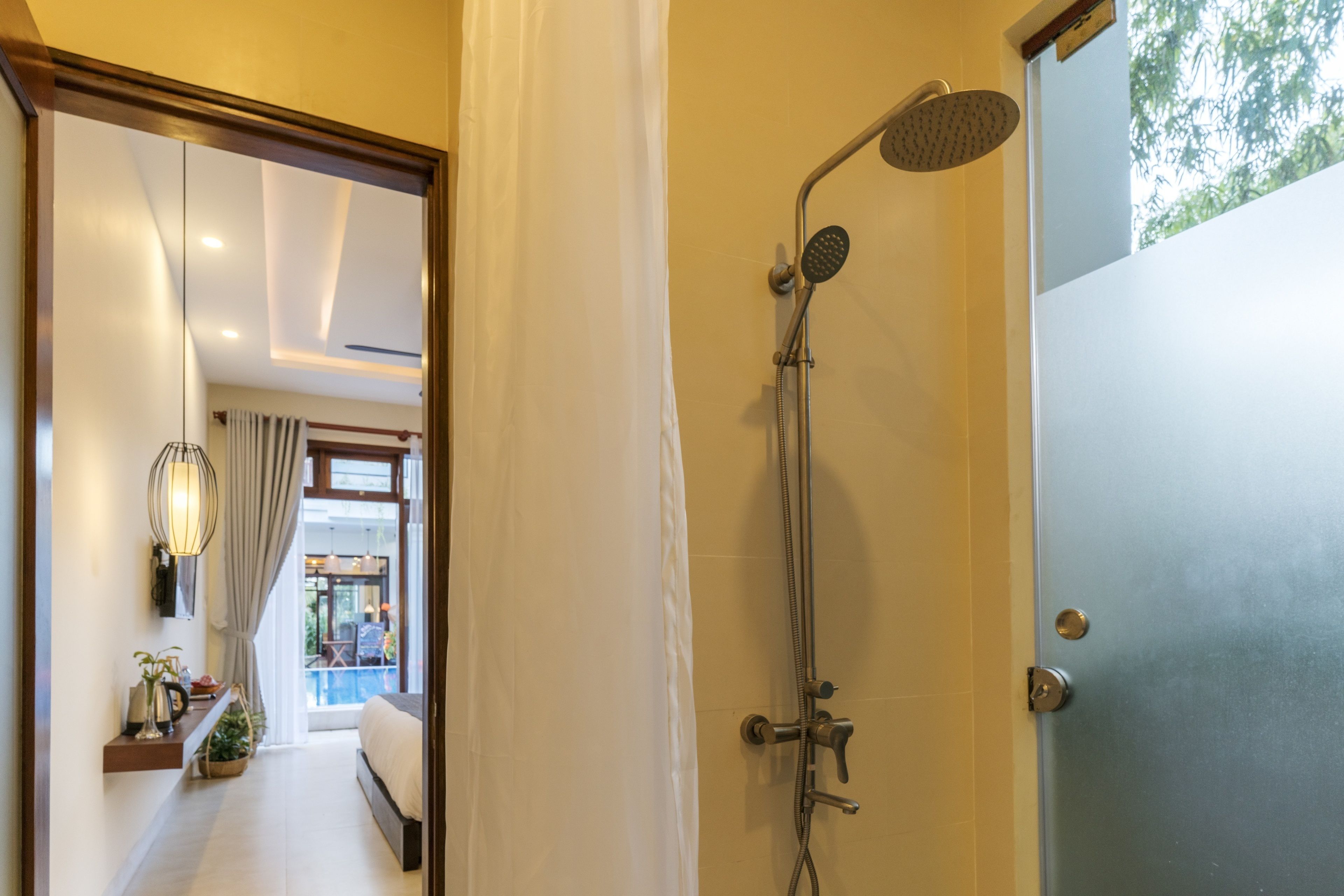 A modern bathroom with a shower, a door, and a view of a balcony with plants and a pool.