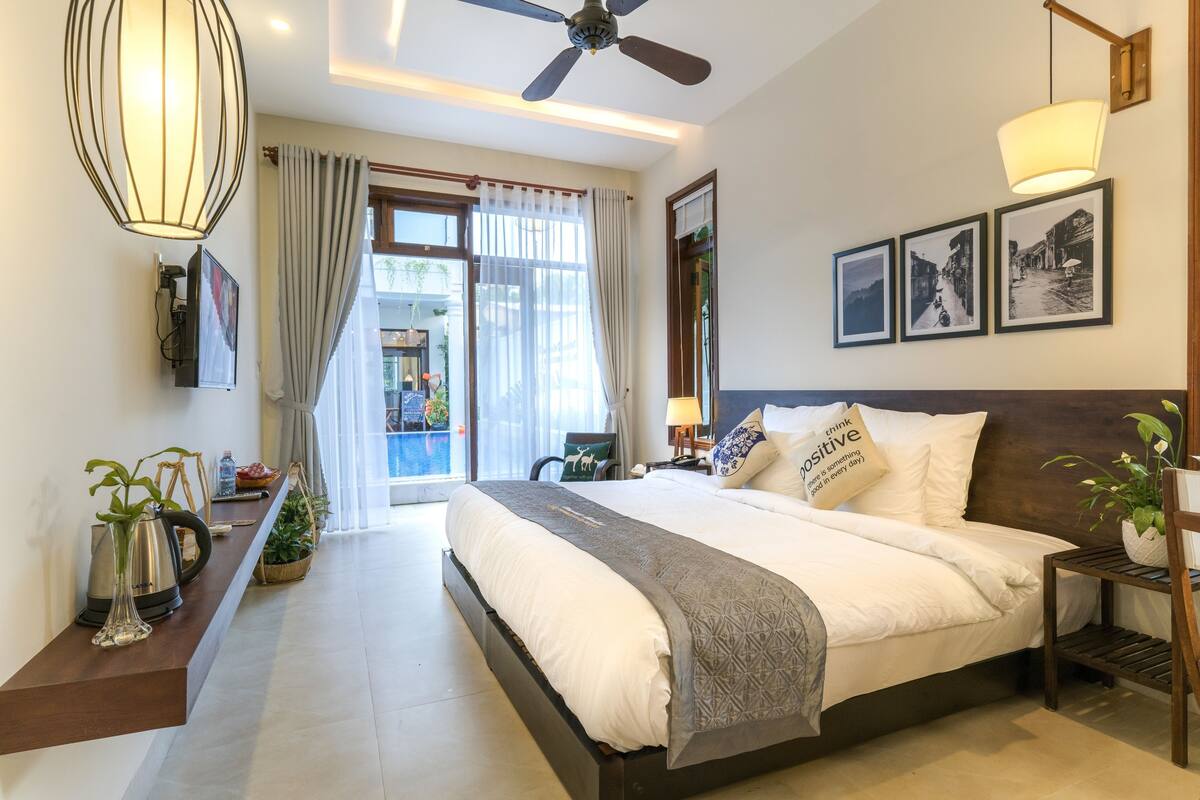 A modern hotel room with a large bed, a ceiling fan, a view of the pool, and a dining area with a table and chairs.