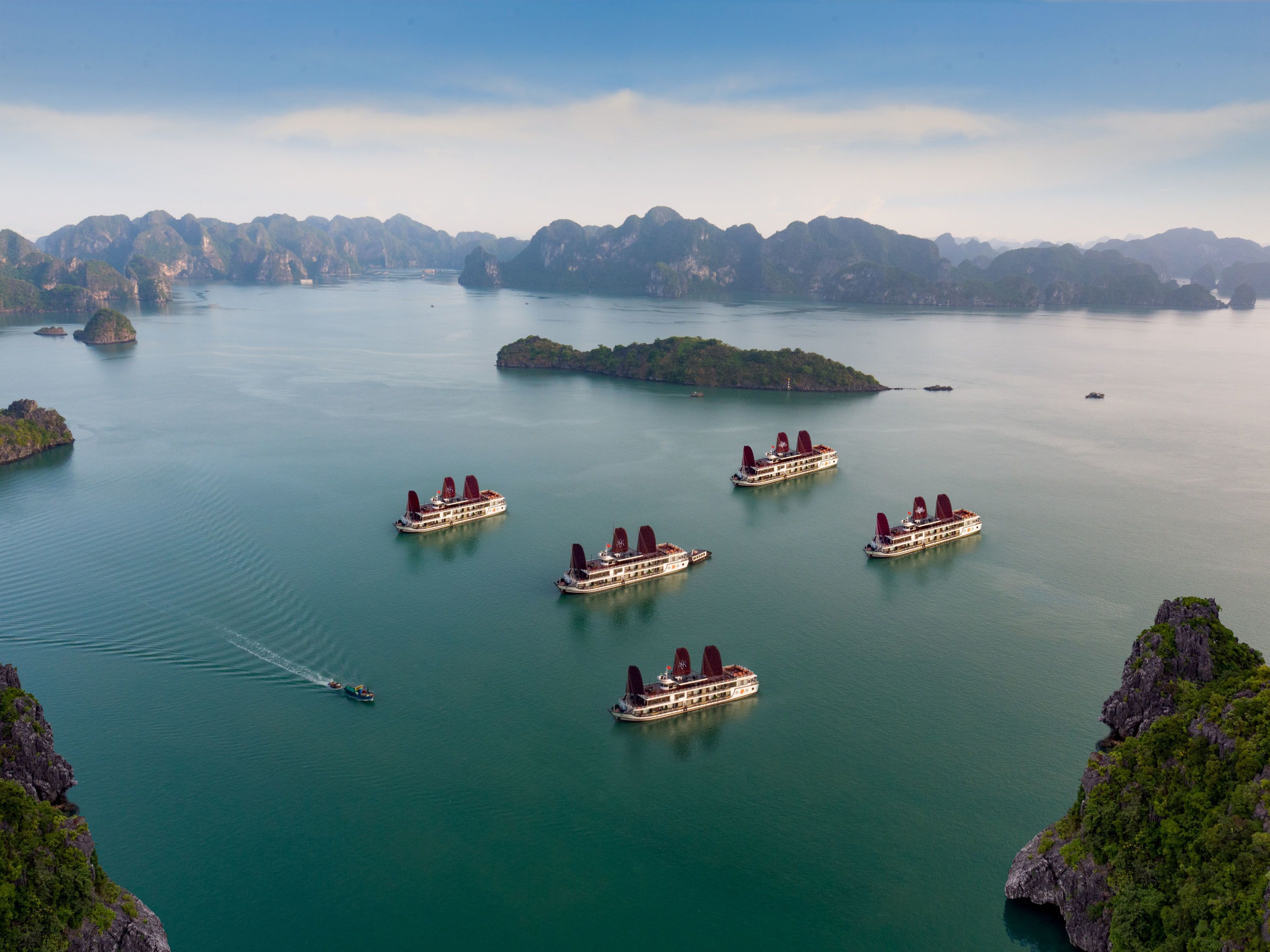 A river cruise with multiple boats, surrounded by lush green islands and rocky cliffs.
