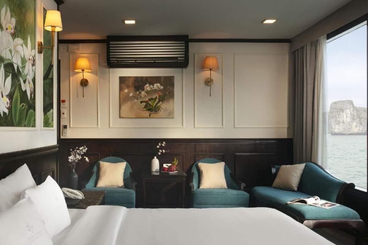 A hotel room with a bed, two armchairs, a small table with a vase, wall-mounted lamps, and a painting.
