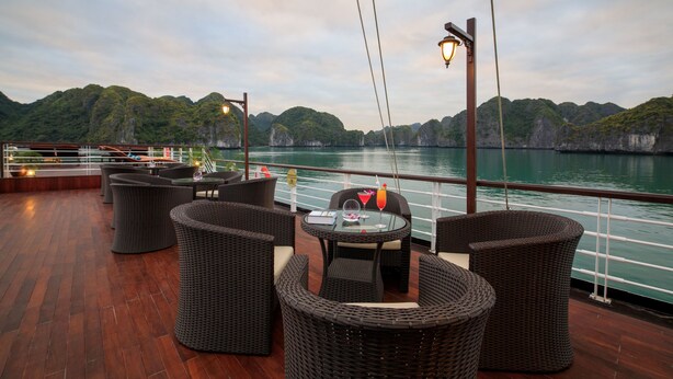 A deck with wicker furniture and a glass table, overlooking a body of water and green hills. - Orchid Trendy Cruise (Tuan Chau)
