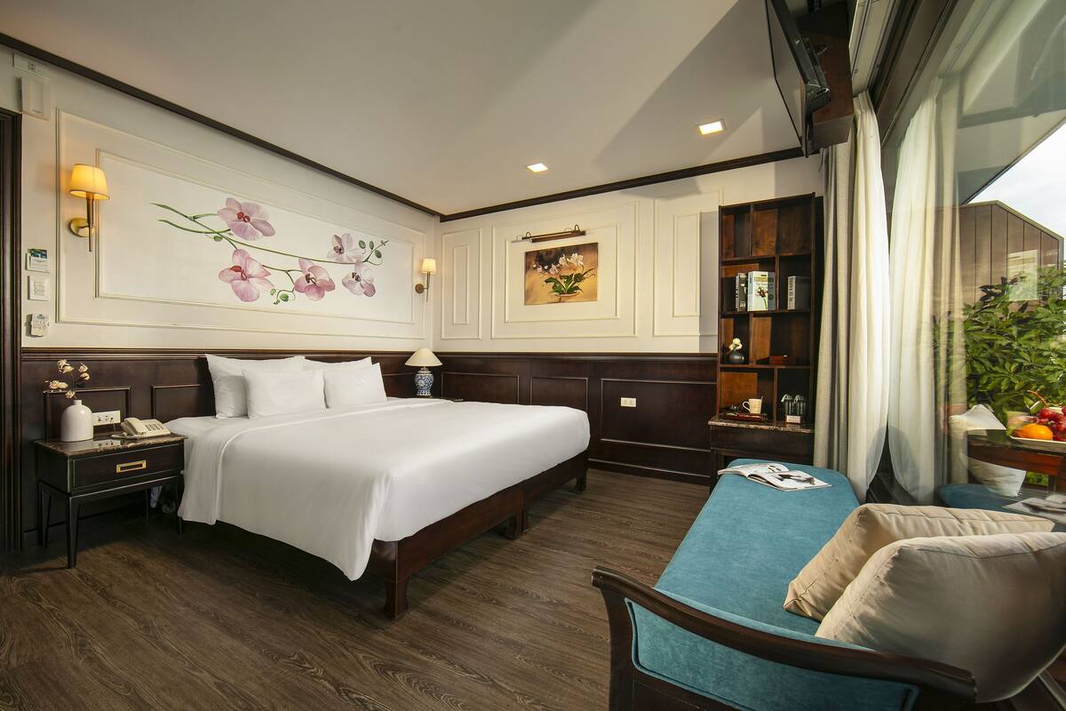 A hotel room with a large bed, bedside tables, a desk with a lamp, a bookshelf, and a window with curtains.