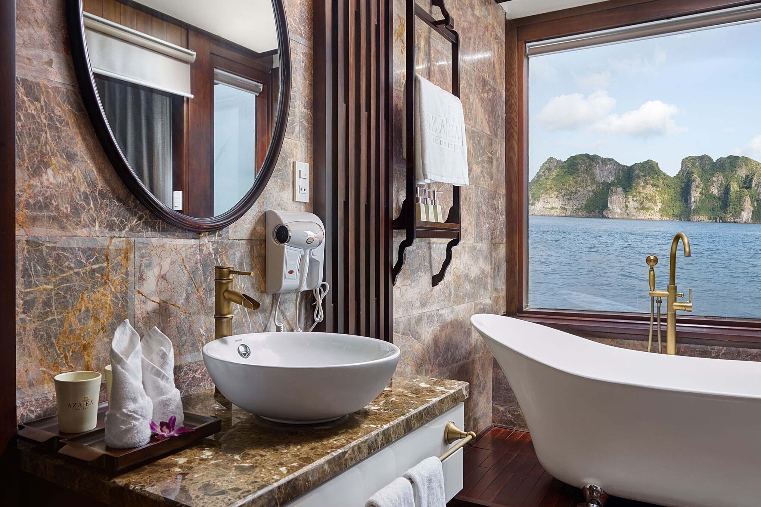 A bathroom with a large oval mirror, a white sink, a freestanding bathtub, and a view of a body of water with cliffs.