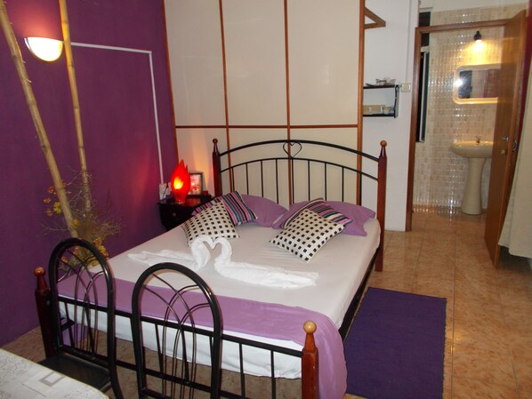 A bedroom with a bed, a nightstand, a lamp, and a bathroom visible through an open door.